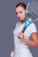 allvarlig konkurrenskraftig. skön ung kvinnor i sporter kläder innehav tennis racket på henne axel och ser på kamera medan stående mot grå bakgrund foto
