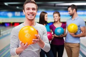 de bäst spelare. stilig ung män innehav en bowling boll medan tre människor kommunicerar mot bowling gränder foto