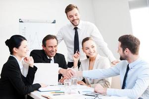 Välkommen på styrelse grupp av självsäker företag människor i formell klädsel Sammanträde på de tabell tillsammans och leende medan två män handskakning foto