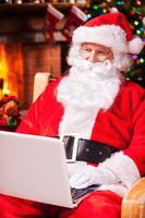 modern santa claus. glad santa claus arbetssätt på bärbar dator och leende medan Sammanträde på hans stol med öppen spis och jul träd i de bakgrund foto