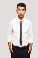 ung och självsäker. stilig ung Afroamerikan man i skjorta och slips ser på kamera och förvaring händer i de fickor medan stående mot grå bakgrund foto