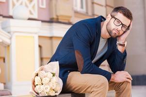 känsla hopplös handla om henne komma. frustrerad ung man i smart jacka lutande hans huvud på hand medan Sammanträde på de bänk med en bukett av blommor om nära honom foto