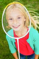 jag am redo till spela topp se av söt liten flicka dölja henne ansikte Bakom badminton racket och leende medan stående på grön gräs foto