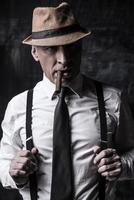självsäker chef. bossig senior man i hatt rökning cigarr och justeras hans hängslen medan stående mot mörk bakgrund foto