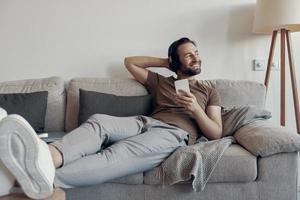 sorglös ung man i hörlurar innehav smart telefon medan avkopplande på de soffa på Hem foto