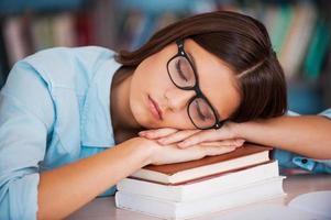 trött av studerar. trött ung kvinnor innehav henne huvud på de bok stack och sovande medan Sammanträde på de bibliotek skrivbord foto