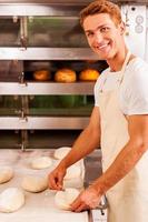 självsäker bagare på arbete. självsäker ung man i förkläde arbetssätt med deg och leende medan stående mot ugn med bröd i den foto