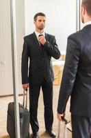 redo för företag resa. självsäker ung man i formell klädsel justeras hans slips medan stående mot spegel i hotell rum foto