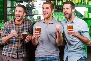 mål tre Lycklig ung män i tillfällig ha på sig innehav glasögon med öl och gestikulerar medan stående på de bar disken och ser bort foto