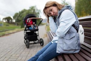 ung kvinna mor av postpartum depression sitter med inbyggt känslor i de parkera på en bänk Nästa till en bebis sittvagn foto