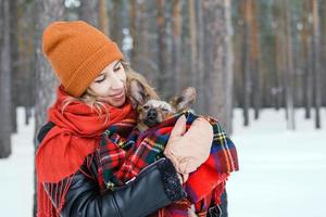 skön ung kvinna är kel liten hund insvept i röd rutig pläd på en promenad i vinter- skog. foto