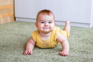 porträtt av leende liten caucasian bebis flicka i gul klänning. barn påfrestande till krypa på en golv. foto