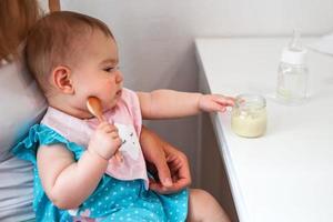 bebis flicka innehar trä- sked i hand och försöker till äta själv vegetabiliska puré från glas burk. foto