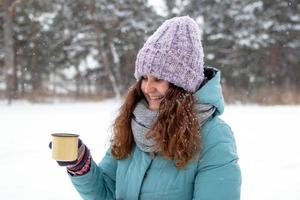 skön leende kvinna med lång lockigt hår i blå vinter- jacka och stickat färgrik handskar är innehav i hand kopp av varm te på en promenad i vinter- snöig skog. foto