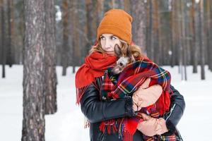 skön kvinna är innehav i händer henne liten hund insvept i röd rutig pläd på en promenad i vinter- skog. foto