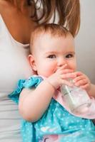 leende liten caucasian bebis flicka i blå klänning dricka vatten från flaska på de mors knäna. barn lär till håll en flaska. foto