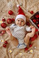 förtjusande leende liten flicka i santa claus röd hatt är om på en beige pläd med röd och vit jul dekorationer och jul lampor och innehav henne tår i händer, topp se. foto