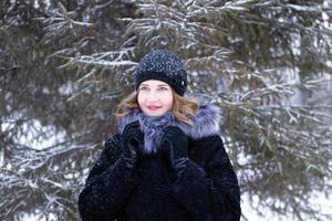 porträtt av skön leende kvinna med kort blond hår i svart falsk päls täcka med grå fluffig huva och svart stickat hatt på en promenad i vinter- barr- skog. foto