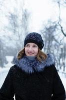 porträtt av ung skön kvinna med kort blond hår i svart falsk päls täcka med grå fluffig huva och svart stickat hatt är gående i vinter- snöig parkera. foto