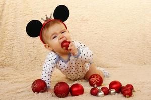 söt liten flicka med mus öron är Sammanträde på en beige pläd och spelar med röd skinande jul bollar. foto