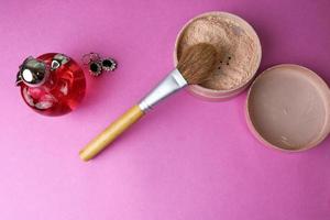 skönhet låda, smulig matt mineral pulver med en särskild naturlig brun trä- borsta från naturlig tupplur för smink, rosa parfym och skinande silver- örhängen på en rosa lila bakgrund. platt lägga. topp se foto