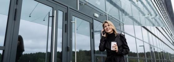 ung kvinna affärsman talande på de telefon med en glas av kaffe på de bakgrund av ett kontor byggnad foto