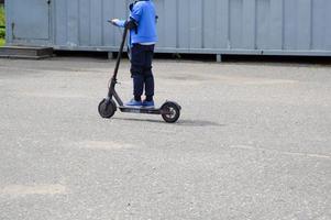 en pojke, en man rider längs de parkera väg på en ny modern elektrisk skoter på batterier, stående på den med två ben foto