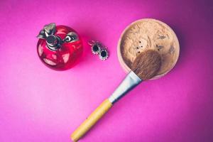 skönhet låda, smulig matt mineral pulver med en särskild naturlig brun trä- borsta från naturlig tupplur för smink, rosa parfym och skinande silver- örhängen på en rosa lila bakgrund. platt lägga. topp se foto