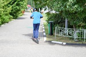 en pojke, en man rider längs de parkera väg på en ny modern elektrisk skoter på batterier, stående på den med två ben foto