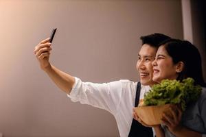 en leende par av manlig och kvinna använder sig av telefon i de kök. tar selfie eller leva strömning via smartphone medan framställning en vegan mat. modern friska livsstil begrepp. matlagning på Hem tillsammans foto