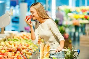 endast de bäst frukt och grönsaker. skön ung kvinna innehav äpple och lukta den med leende medan stående i en mat Lagra foto