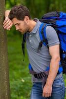 trött resande. sida se av trött ung man med ryggsäck lutande på de träd och förvaring ögon stängd foto