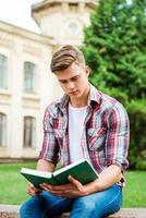 stilig bokmal. självsäker manlig studerande läsning bok medan Sammanträde på de bänk och i främre av universitet byggnad foto