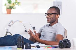 känsla självsäker och avslappnad. stilig ung afrikansk man i tillfällig ha på sig Sammanträde på hans arbetssätt plats och arbetssätt på digital läsplatta foto