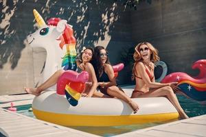 attraktiv ung kvinnor i badkläder leende medan flytande på uppblåsbar enhörning i simning slå samman utomhus foto