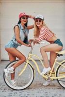 njuter Bra tid tillsammans. sida se av leende ung kvinna Sammanträde på cykel medan henne kvinna vän stående i främre av den foto