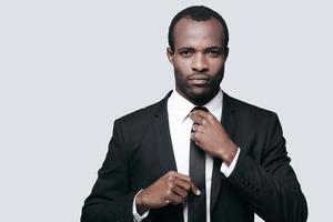 perfekt affärsman. porträtt av självsäker ung afrikansk man justeras hans slips medan stående mot grå bakgrund foto
