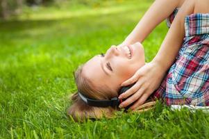 njuter musik i natur. sida se av skön ung kvinna i hörlurar lyssnande till de musik och leende medan liggande på de grön gräs foto