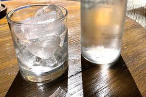 selektiv fokus, en klar glas fylld med is och klar glas flaska innehar kall mineral vatten sitter på trä- tabell foto