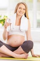 rena vatten är Viktig för min bebis. skön gravid kvinna innehav en flaska med vatten medan Sammanträde i lotus placera foto