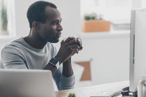 njuter morgon- kaffe. omtänksam ung afrikansk man innehav en kopp och ser på de dator övervaka medan Sammanträde på de skrivbord i kreativ kontor foto
