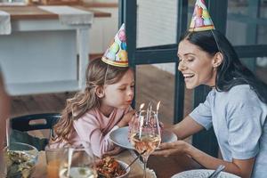 Lycklig liten flicka blåser ljus på födelsedag kaka medan har middag med familj foto