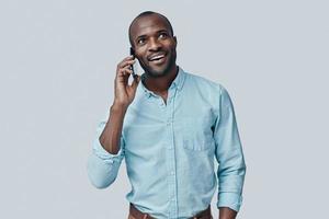 stilig ung afrikansk man talande på de smart telefon och leende medan stående mot grå bakgrund foto