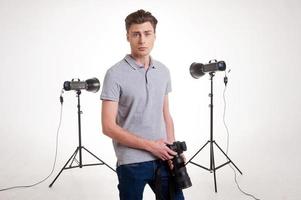 självsäker fotograf. stilig ung man i polo skjorta lutande på de stativ med kamera medan stående i studio med belysning Utrustning på bakgrund foto