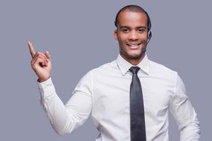 din text här. självsäker ung afrikansk man i headsetet pekande bort och leende medan stående mot grå bakgrund foto