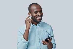 stilig ung afrikansk man lyssnande musik använder sig av smart telefon och leende medan stående mot grå bakgrund foto