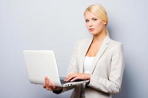 stödjande din företag. självsäker ung affärskvinna innehav bärbar dator medan stående mot grå bakgrund foto