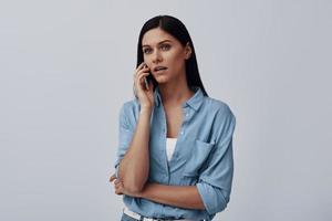 attraktiv ung kvinna talande på de smart telefon medan stående mot grå bakgrund foto