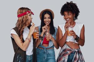 tre attraktiv ung kvinnor dricka uppfriskande cocktail och leende medan stående mot grå bakgrund foto