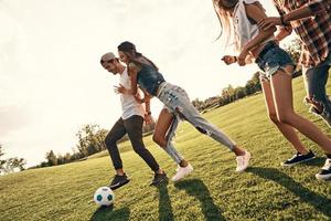 utgifterna bra tid med vänner. grupp av ung leende människor i tillfällig ha på sig njuter trevlig sommar dag medan spelar fotboll utomhus foto